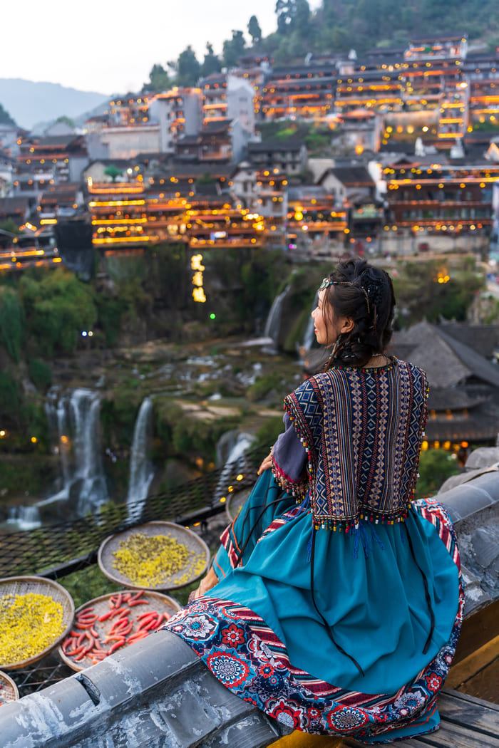 Una mujer vestida con un traje tradicional sentada sobre una barandilla de tejado en la antigua ciudad china de Fenghuang.