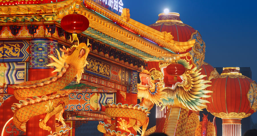 Detalle arquitectónico de un templo chino iluminado por la noche, decorado con intrincadas figuras de dragones dorados y farolillos rojos tradicionales.