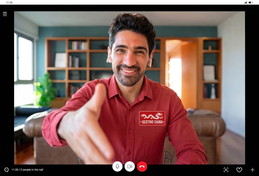 Hombre sonriente en videollamada con camisa roja de "Destino China" ofreciendo la mano a la cámara.
